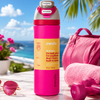 A bright pink Owala water bottle with a yellow label stands on a marble surface outdoors by the beach, with a pink towel, pink bag, sunglasses, and dumbbells nearby.