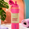 A Owala insulated water bottle with a built-in straw and leak-proof lid, styled on a tabletop with a rolled pink towel, sunglasses, earbuds case, and plant leaves in a bright studio setting.