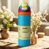 A Owala insulated water bottle with a bright blue body, orange and purple accents, and a yellow flip-top lid, standing on a wooden table with blurred flowers in the background.