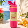 A pink Owala insulated water bottle with a matching pink flip-top lid and a beige wrap label, styled on a sunlit stone surface with a beach and palm leaves in the background, alongside citrus slices and a yellow towel.