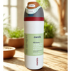 A Owala insulated water bottle with a bright blue body, orange and purple accents, and a yellow flip-top lid, standing on a wooden table with blurred flowers in the background.