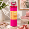 A bright pink Owala insulated water bottle with a gray lid and orange label, styled on a marble coaster in a warm, sunlit room with books, a plant, and a candle in the background.