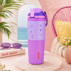 A Lavender Owala water bottle with a matching purple and pink flip-top lid sits on a round stone coaster in a styled beach-themed setting with sunglasses, a book, and tropical decor.