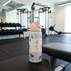 A Owala water bottle with a light blue body, peach lid, and colorful food illustrations, sitting on a gym bench with a blurred gym background.