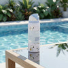 A light blue and white Owala water bottle with Snoopy illustrations stands on a glass table next to a swimming pool with a blurred background of green plants.