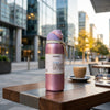 A pink Owala insulated water bottle with a white label stands on a wooden outdoor cafe table, with a coffee cup, sunglasses, and blurred city buildings in the background.