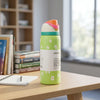 A light green Owala water bottle with white daisy patterns and a white paper label, sitting on a wooden table in a bright, modern room with books and a window in the background.