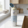 A white insulated water bottle with a light blue label and cartoon bunny illustrations, shown on a kitchen counter with its lid open. The background is a softly blurred kitchen interior with light wood cabinets and a bright window.