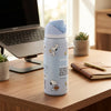 A light blue Owala water bottle with Snoopy illustrations sits on a wooden desk in a bright office setting, next to a laptop, notebook, and small potted succulents.