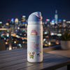 A white Owala insulated water bottle with a light blue lid and pink accents, featuring cute cartoon characters and a quote, set on a wooden table with a blurred city skyline at night in the background.