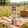A reusable insulated water bottle with a pink lid and a white label featuring red tulips, standing on a picnic blanket in a grassy field with a wicker basket in the background.