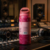 A pink water bottle with a floral pattern and a white label stands on a mixing board in a recording studio setting with speakers and headphones visible in the background.