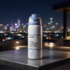An Owala insulated water bottle with a light blue flip-top lid stands on a dark metal table, with a blurred nighttime city skyline in the background.