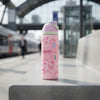 A pink Owala Hello Kitty insulated water bottle with a matching pink lid, standing on a concrete ledge in an outdoor transit station with blurred trains and tracks in the background.