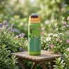 A green water bottle with Minecraft-themed graphics stands on a wooden table in a garden setting with flowers and greenery in the background.