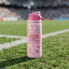 A pink Owala Hello Kitty water bottle with a free-sip lid stands on green grass in a stadium, with blurred stands in the background.