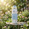 A white insulated water bottle with a light blue flip-top lid stands on a small round table in a sunlit garden setting with flowers and greenery in the background.