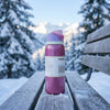 A pink and purple Owala water bottle with a white label sits on a snow-dusted wooden bench in a snowy outdoor setting with trees and mountains in the background.