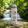 A pink Owala insulated water bottle with a blue and pink flip-top lid stands on a mossy rock in front of a blurred forest stream and waterfall background.