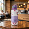 A white Owala insulated water bottle with purple and gray lid details, featuring cartoon character graphics and a purple label, sitting on a wooden table in a blurred cafe setting.