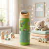A green children's water bottle with Minecraft-themed character graphics stands on a wooden table in a softly styled nursery setting. A teddy bear rests on folded pastel blankets nearby, with toys and warm string lights in the background.