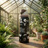 A Owala insulated water bottle with a black body, white graphic illustrations, and a brown lid, standing on a wooden table inside a sunlit greenhouse surrounded by green plants.