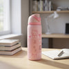 A pink Hello Kitty insulated water bottle with a matching pink lid and strap, standing on a light wood desk in a bright room with books and a notebook nearby.