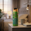 A green insulated water bottle with Minecraft-themed graphics stands on a bedside table in a softly lit bedroom. A gray mug, book, and wall lamp are visible in the background.