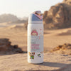 A white Owala insulated water bottle with cute cartoon illustrations and a light blue lid, standing on a sandy rock with a blurred desert landscape in the background.