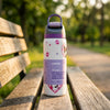 A white Owala water bottle with purple accents and cartoon graphics sits on a wooden park bench with a blurred green outdoor background.