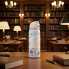 A light blue insulated water bottle with a pink lid and white label stands on a wooden table in a cozy, blurred library setting with bookshelves, lamps, and books.
