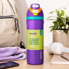 A purple Owala insulated water bottle with a bright green label and a teal-and-orange flip-top lid, standing on a wooden table in a styled room setting with a phone, towel, headphones, notebook, and plant in the background.