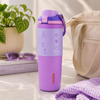 A Lavender Owala insulated water bottle with a matching purple lid and carry loop, styled on a light marble surface with a beige tote bag, sunglasses, a book, and a knit blanket in the background.