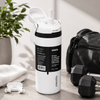 A white Owala insulated water bottle with a black label and flip-top lid, styled on a tabletop with a towel, earbuds case, dumbbells, and a black bag in the background.