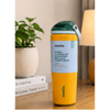 A yellow Owala water bottle with a dark green lid and a light blue wrap label, standing on a wooden table next to a potted plant and a lamp.