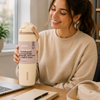 A woman in a beige sweatshirt holds an Owala leak-proof water bottle with a lavender label at a wooden desk, with a laptop, notebook, and pen nearby.