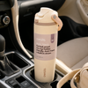 An Owala insulated water bottle with a built-in straw, placed in a car cup holder. The bottle is cream-colored with a lavender label and a light wood-grain carrying loop.