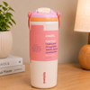 An Owala insulated water bottle with a pink and white design, featuring a pink label with text and a pink flip-top lid, set on a wooden table next to a potted plant and a lamp.