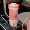 A Owala insulated water bottle with a pink and cream design, resting in a car cup holder next to a matching pink handbag.