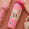A pink Owala water bottle with a red carry loop and beige label, styled on a peach textured surface with a pink dumbbell, towel, and white headphones.