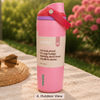 A pink Owala insulated water bottle with a flip-top lid and carrying loop, shown outdoors on a stone surface with blurred greenery and pink flowers in the background. A straw hat rests nearby, and the label on the bottle reads 'owala' with product copy.