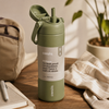 An Owala insulated water bottle in Olive green, shown on a wooden table with a beige backpack, notebook, and plant in the background.