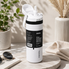 A white Owala insulated water bottle with a black label and flip-top lid, styled on a stone surface with plants, sunglasses, and a folded towel in a bright outdoor setting.