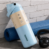 A light blue Owla water bottle with a beige label, leaning against a rolled blue yoga mat next to a black kettlebell on a gray concrete floor against a white brick wall.