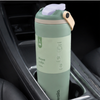 A mint green insulated water bottle with a light purple flip-top lid and beige carrying loop, placed in a car cup holder. The bottle has a white paper sleeve with product details and the brand name 'wala.